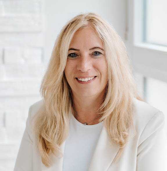 Porträt von Anke Brettschneider, Systemisch-heilpädagogische Beraterin, aus Neuendettelsau. Mit langen blonden Haaren, einem weißen Blazer vor hellem Hintergrund.
